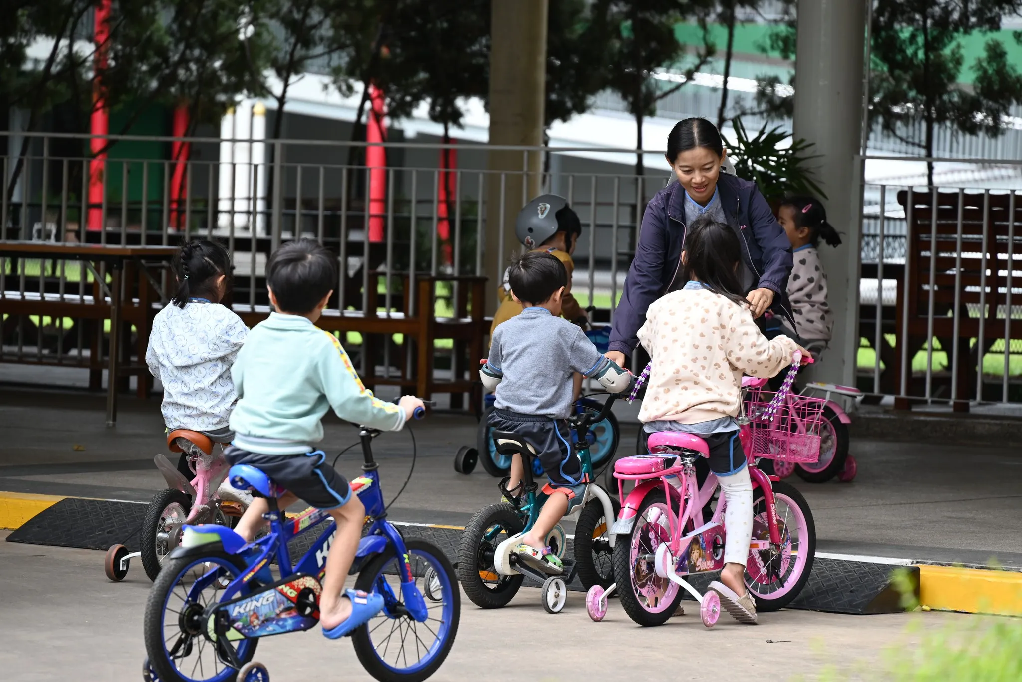 เด็กเล็กหลายคนกำลังขี่จักรยานคันเล็กและจักรยานทรงตัวอยู่ในพื้นที่เล่นกลางแจ้งที่มีหลังคาคลุม มีครูหรือผู้ดูแลในชุดแจ็คเก็ตสีเข้มยืนอยู่ตรงกลางเพื่อช่วยเหลือเด็กหญิงตัวน้อยบนจักรยานสีชมพู เด็กๆ สวมชุดนักเรียนลำลอง และพื้นหลังเผยให้เห็นบริเวณสนามเด็กเล่นที่มีรั้วกั้นและต้นไม้