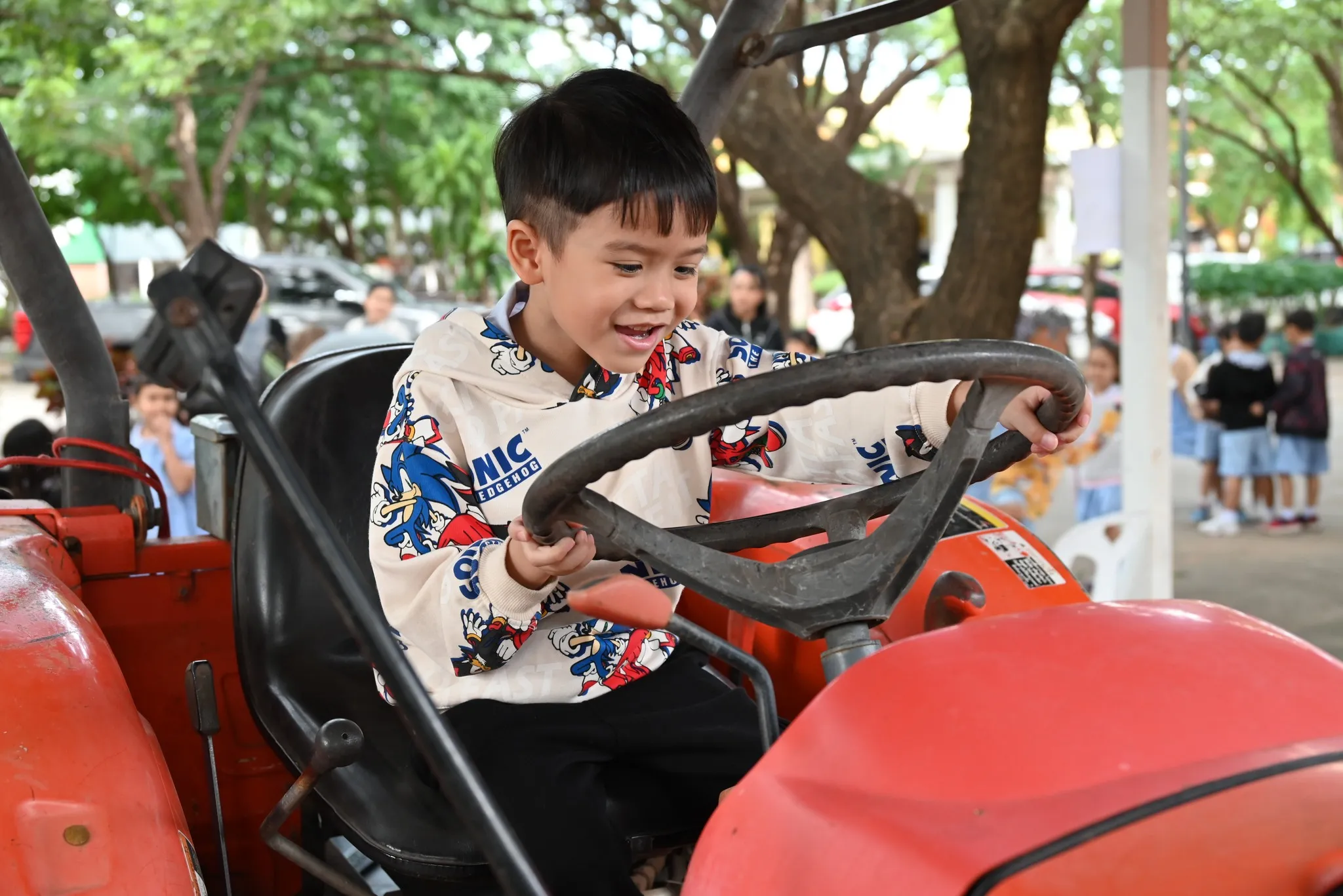 ภาพถ่ายระยะใกล้ของเด็กชายที่มีสีหน้ายิ้มแย้มแจ่มใส นั่งอยู่บนที่นั่งคนขับของรถแทรกเตอร์สีแดงที่จอดอยู่ เขาสวมเสื้อฮู้ดสีครีมลายการ์ตูน 'Sonic the Hedgehog' และวางมือทั้งสองข้างบนพวงมาลัยสีดำขนาดใหญ่เพื่อสมมติว่ากำลังขับรถอยู่ พื้นหลังเป็นพื้นที่กลางแจ้งที่มีแสงแดดส่องถึง พร้อมต้นไม้และเด็กคนอื่นๆ